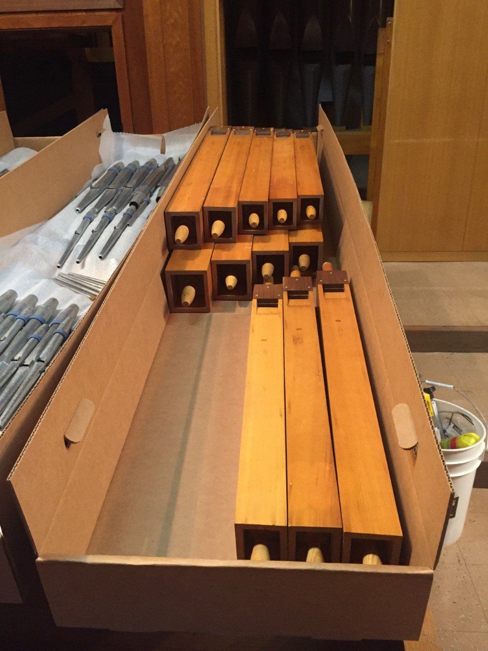 Open cardboard box holding wooden bar clamps and blue-handled tools on a tabletop.