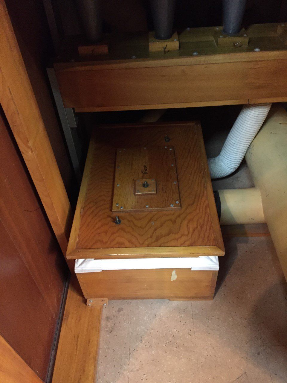 Wooden crawlspace hatch with a small access door under floor joists and a white vent hose nearby