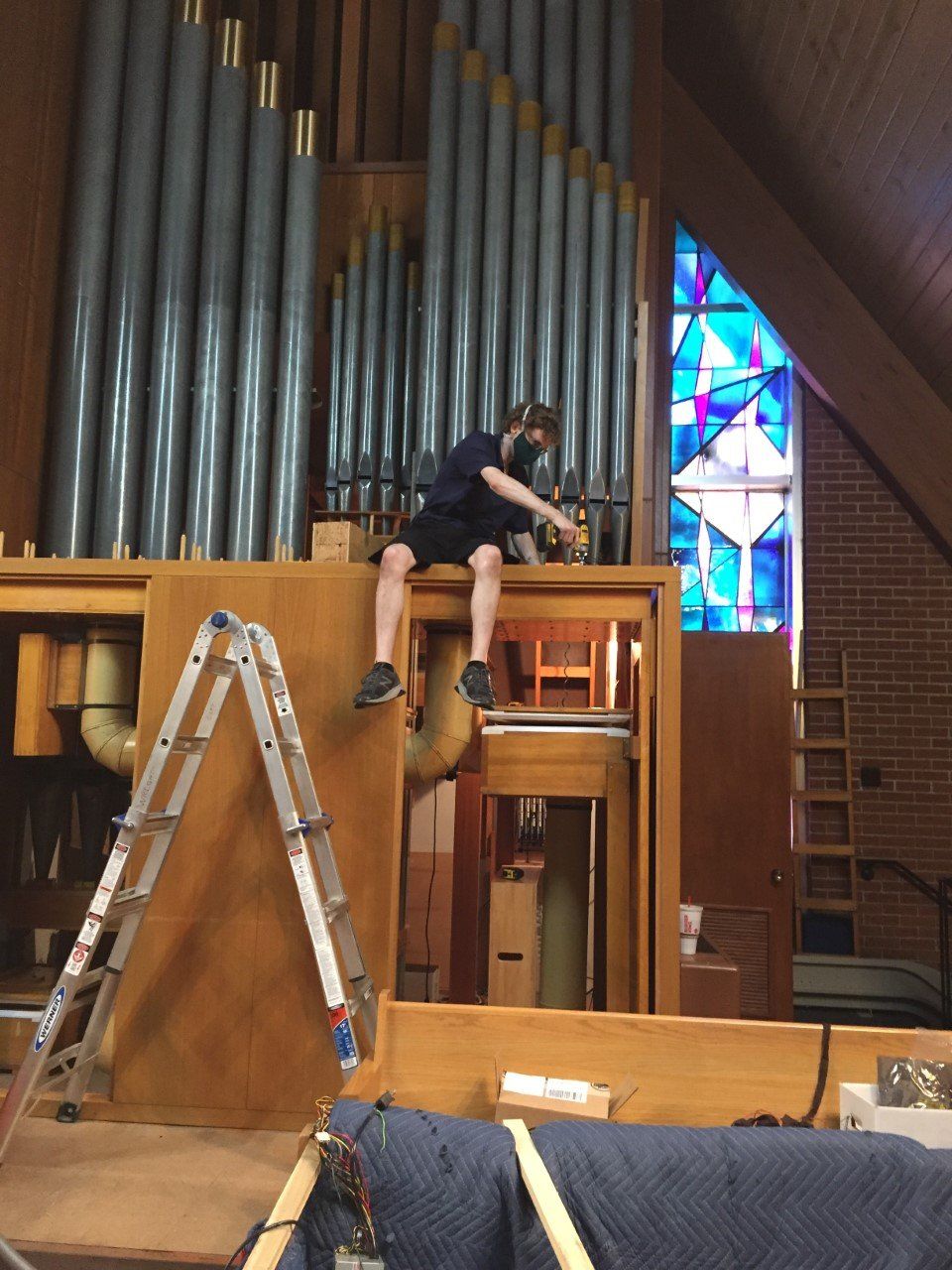Person working on a large organ in a church, with a ladder nearby and stained glass windows in the background