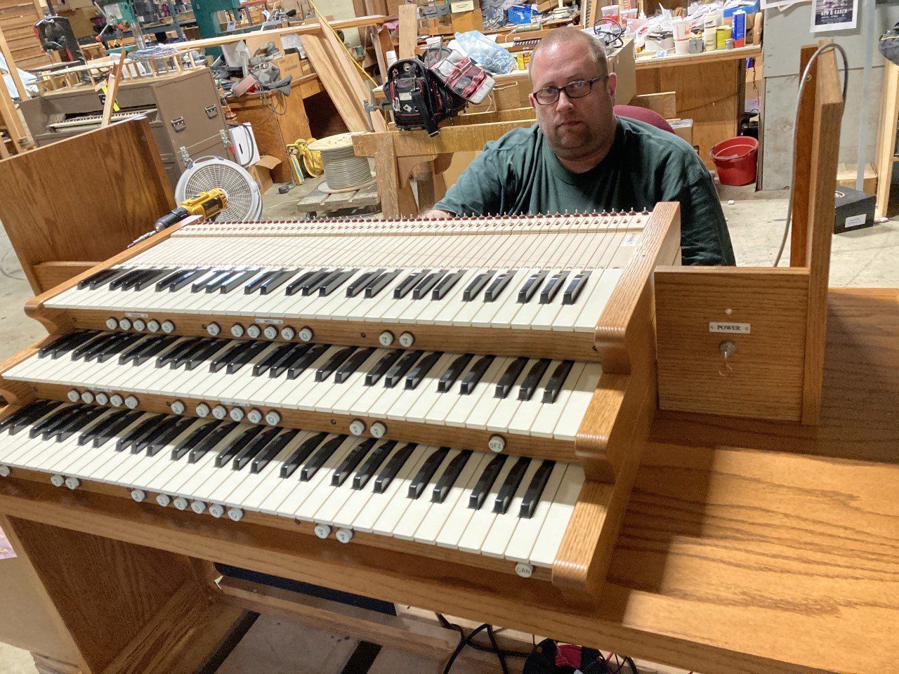 Man playing a large pipe organ in a workshop, with dual keyboards and wooden console.