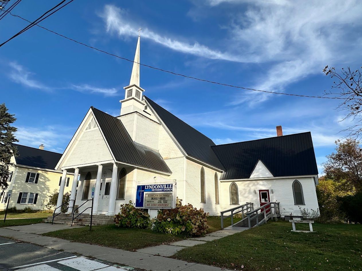 White church with steeple and dark roof, sign, and small ramp on a sunny day.