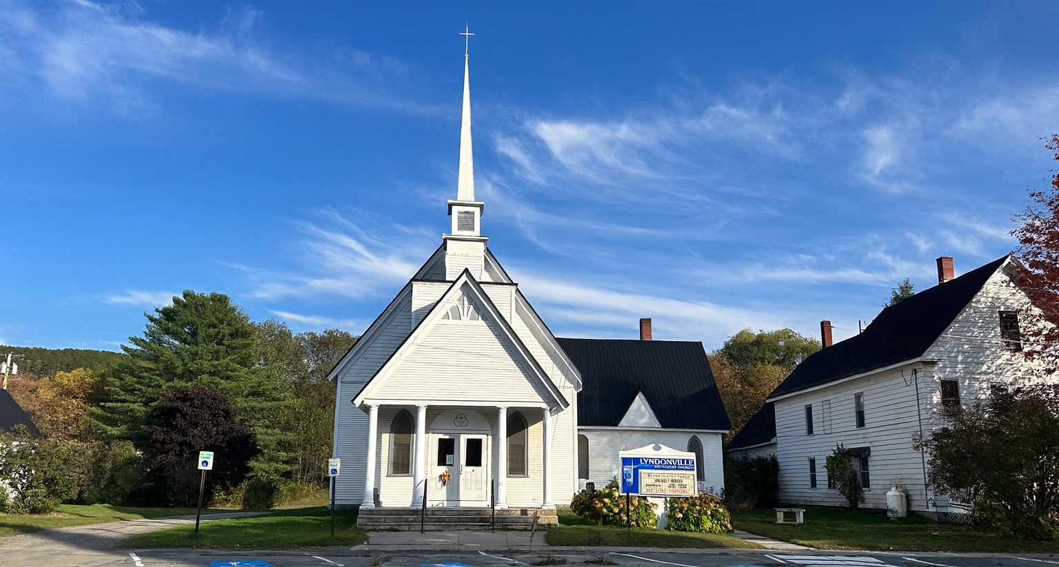White clapboard church with steeple, blue sky background, autumn trees and building to the right.