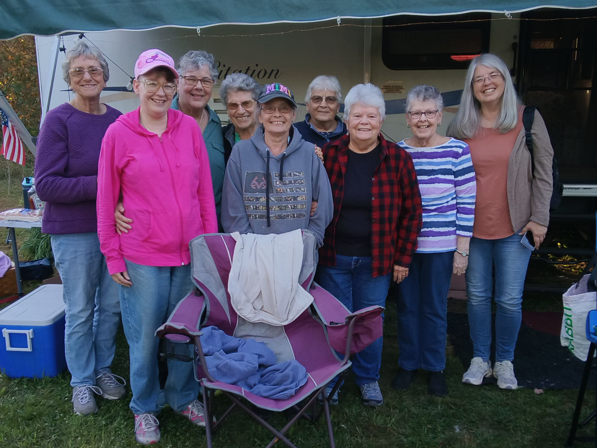 Group of women smiling, posing in front of RV. Some wear hats, sweaters. Outdoors on grass.