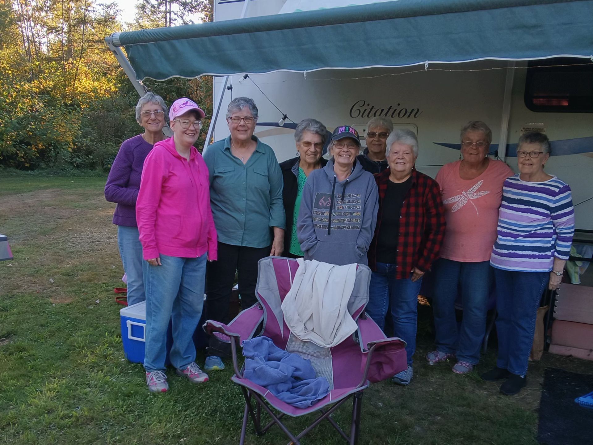 Group of people standing near a camper with an awning. They are smiling, with various colored clothing. Outdoors.