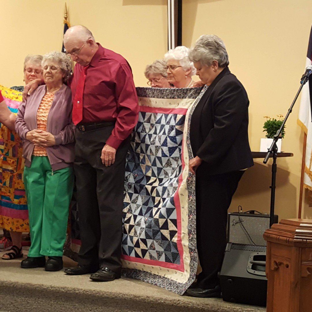 People on stage displaying quilts, one man and several women. The setting is indoors, with a stage.
