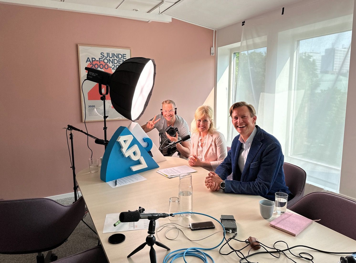 Three people at a table in a studio, recording. One gives a thumbs up. A large light illuminates them.
