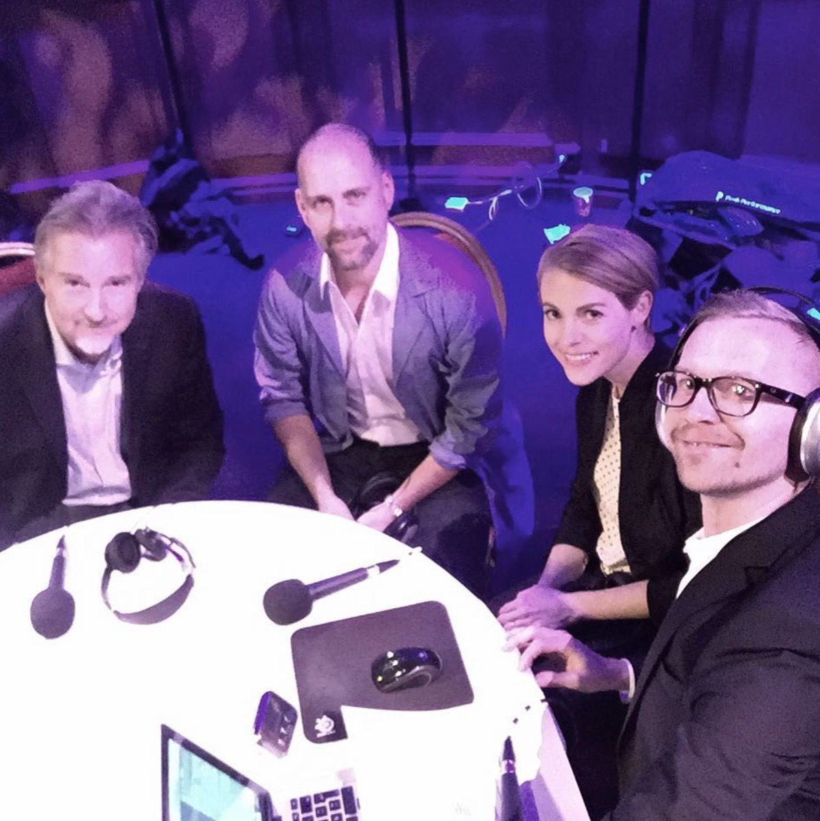 Four people seated around a round table with microphones and equipment, appearing to record a podcast in a dimly lit studio.