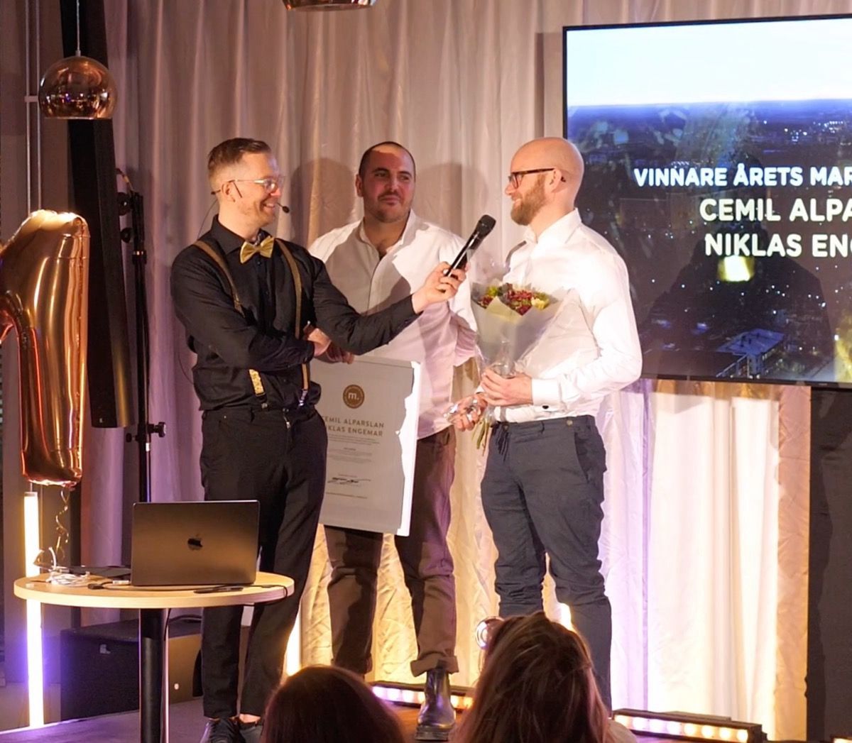 Men on stage, award ceremony. A man holds flowers, another holds a certificate, and a third holds a microphone. Background display.