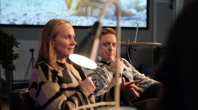 Woman in black/tan patterned jacket and man with mic, talking on a couch, indoors. Bright light in foreground.