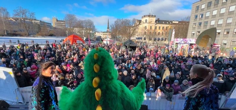 A green dinosaur costume stands before a large crowd on a sunny day. Performers wear sequined outfits.