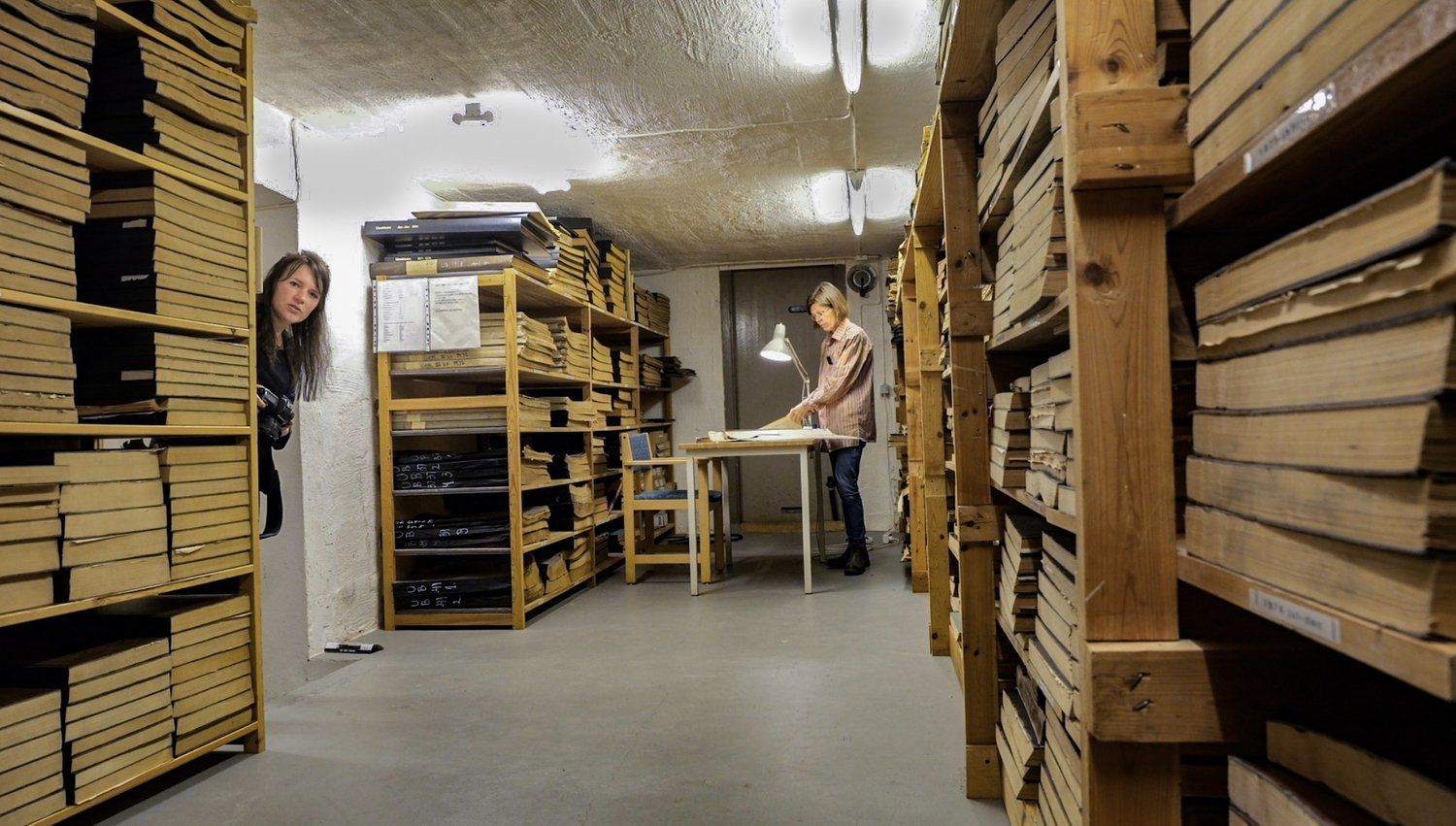 Two people in a library archive. One looks at the camera, the other stands at a table. Wooden shelves hold books.