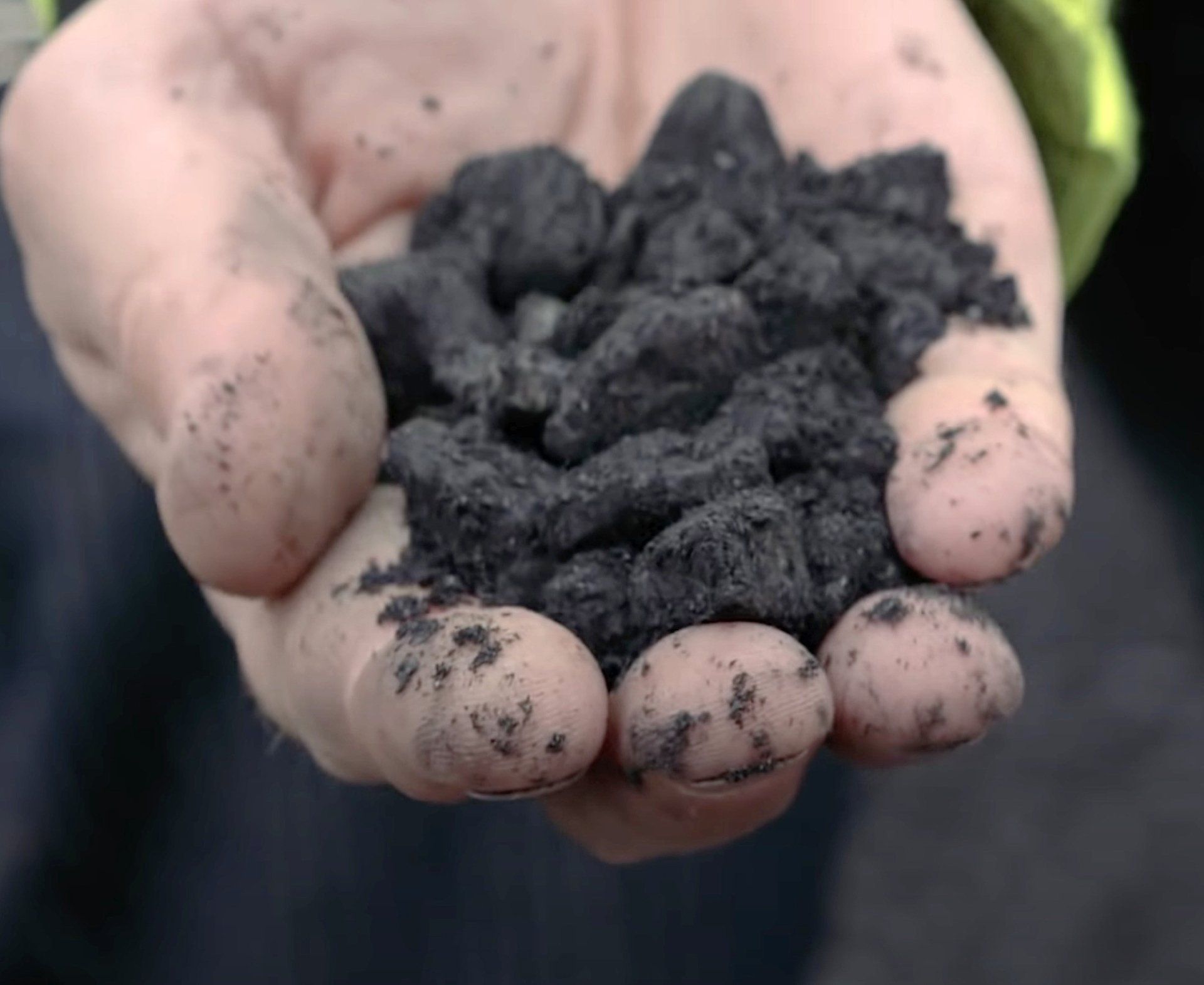 Hand holding a pile of dark, irregularly shaped, crumbly material; likely a product or substance.