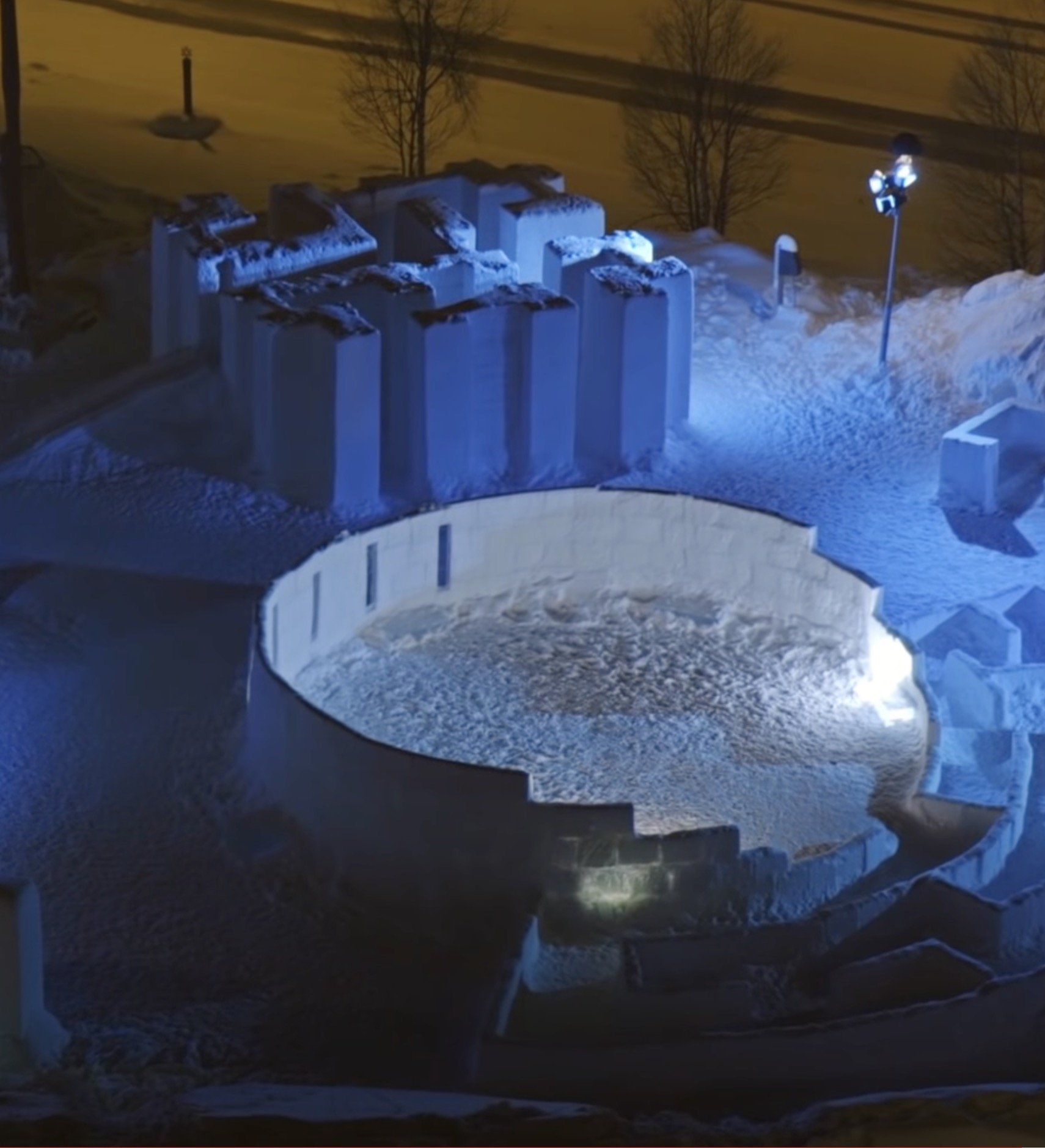 Snow sculpture illuminated at night, featuring a circular structure and surrounding rectangular forms, covered in snow.
