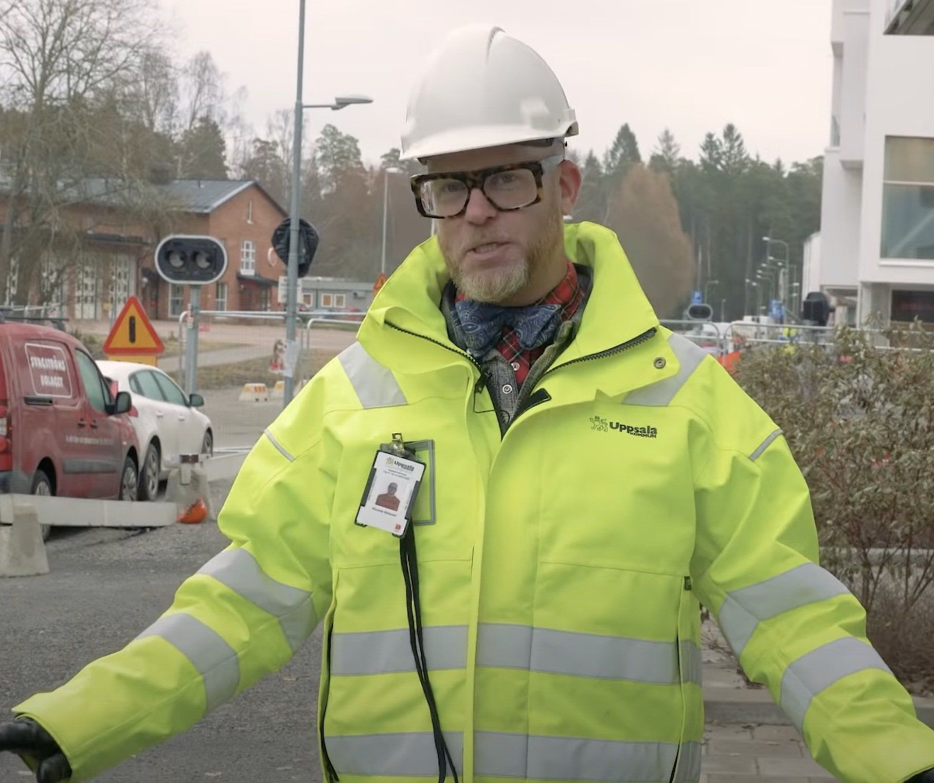 Man in a yellow reflective jacket and hard hat stands on a construction site, gesturing with his hands.