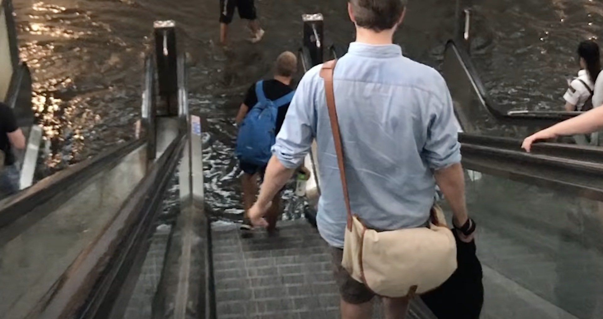 People riding an escalator downwards. A person in a blue shirt with a satchel walks. Dimly lit.