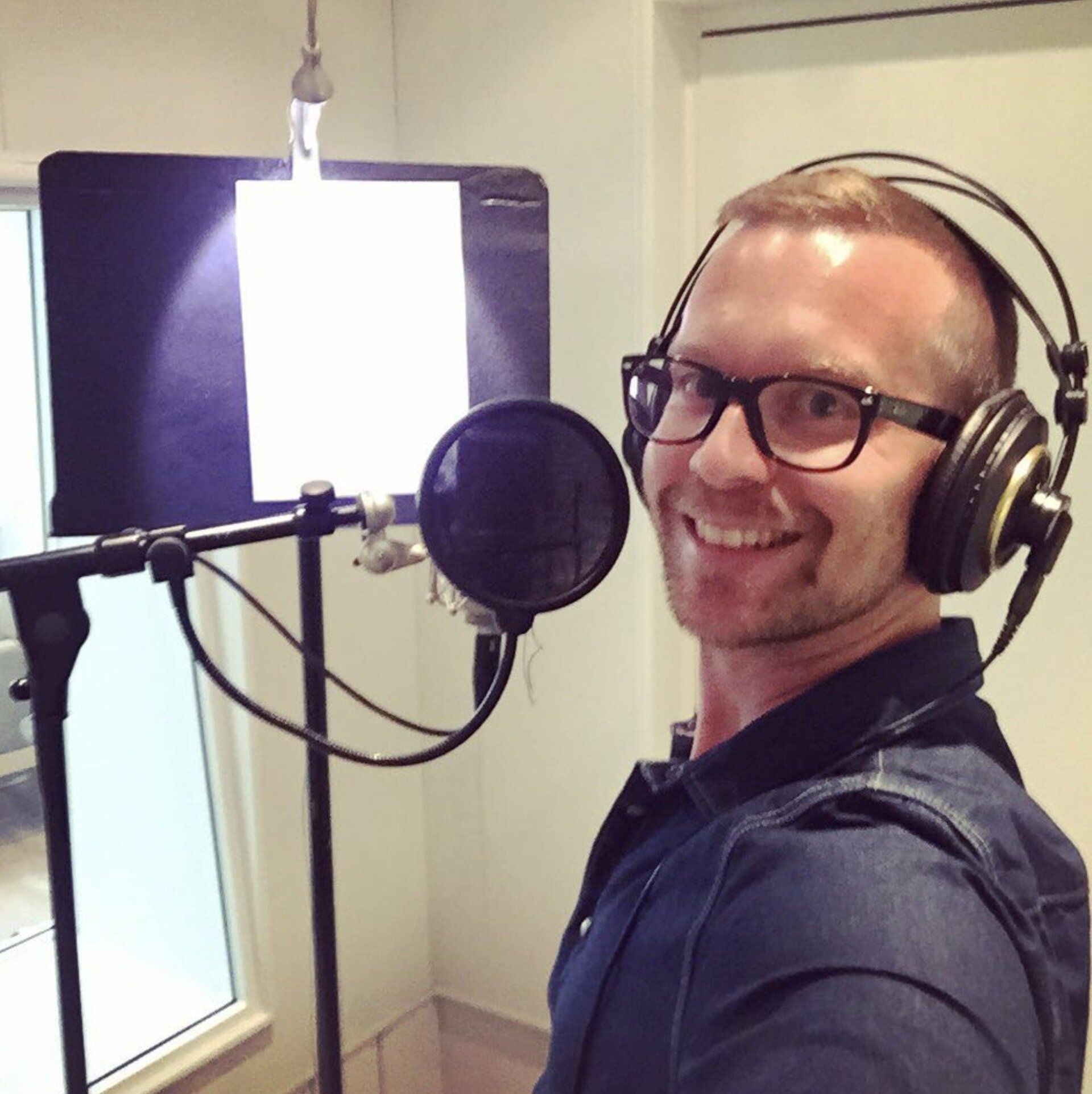 Man with glasses and headphones smiling into a microphone in a recording studio.