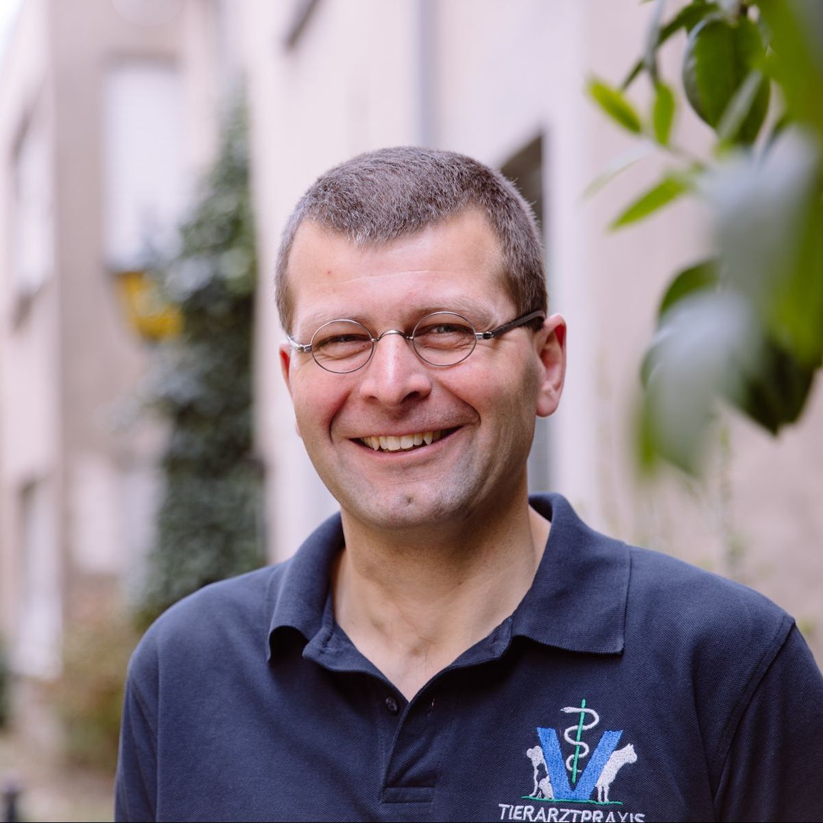 Ein Mann mit Brille lächelt und trägt ein dunkelblaues Poloshirt mit dem Logo einer Tierarztpraxis.