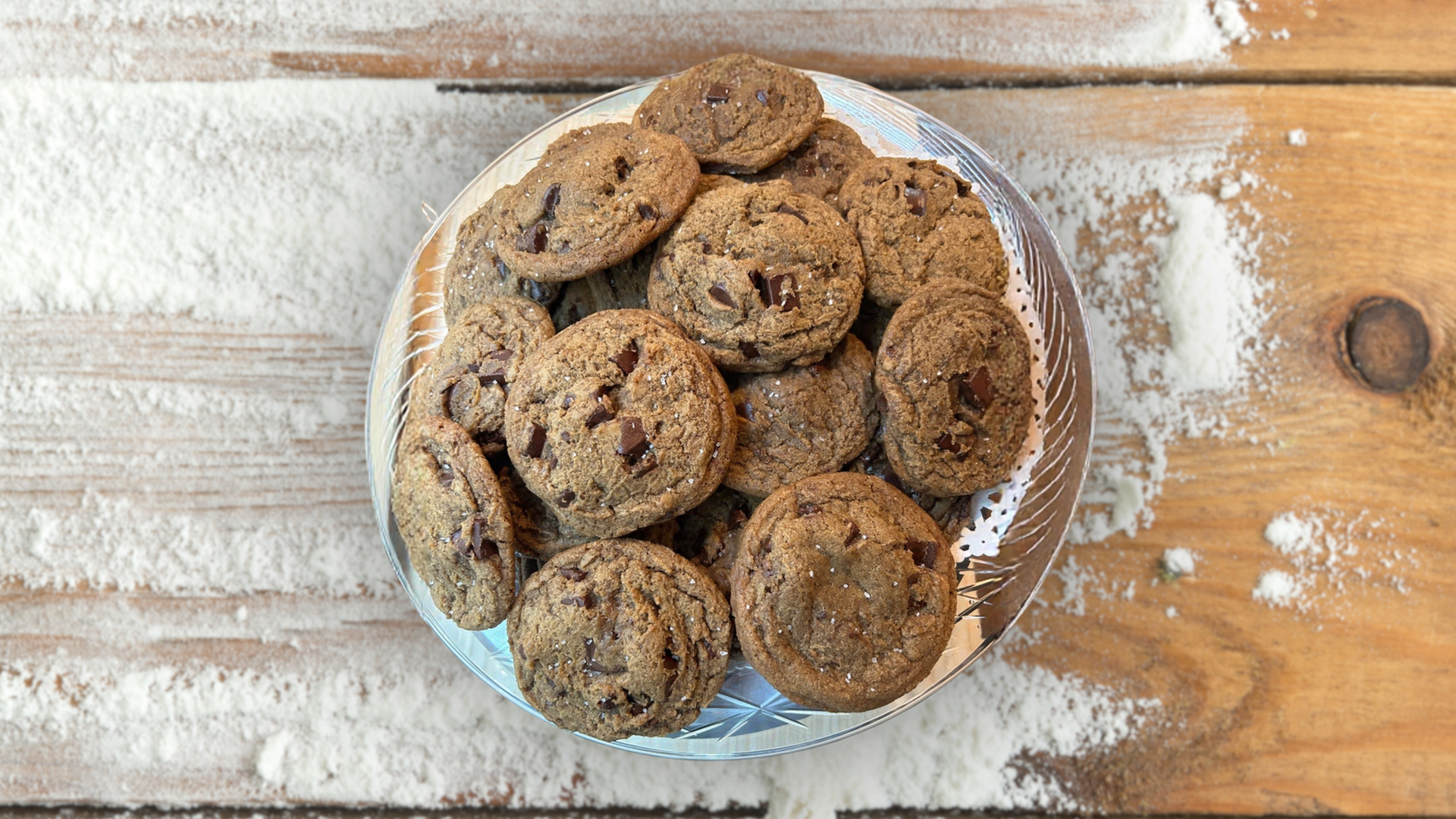 Chocolate chip cookies on a plate, surrounded by spilled flour on a wooden surface.