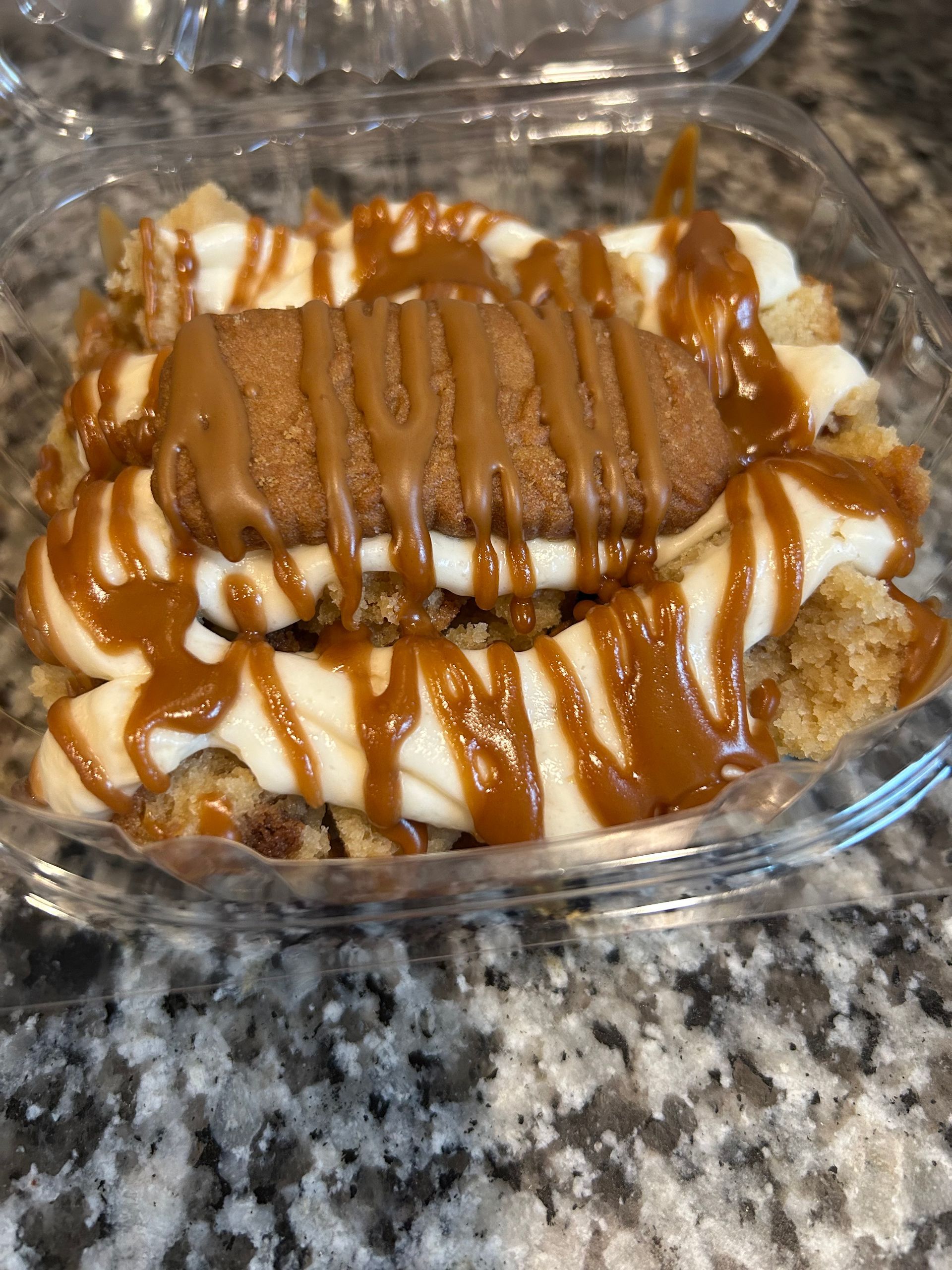 Dessert in plastic container: layers of cake, white cream, and caramel drizzle, topped with a cookie.