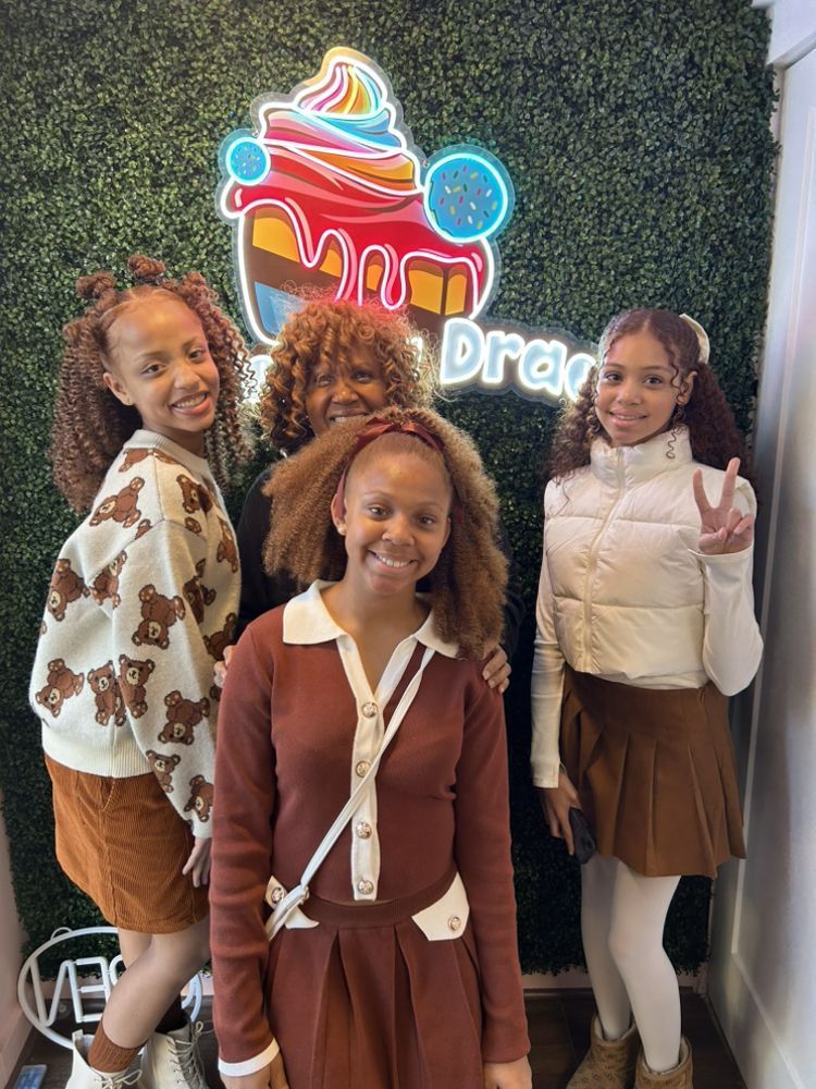 Four people pose in front of a neon sign. Two wear brown, one makes a peace sign. Green backdrop.