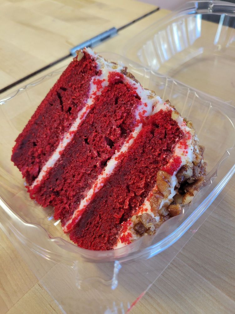 Slice of red velvet cake with white frosting and pecans in a clear plastic container.