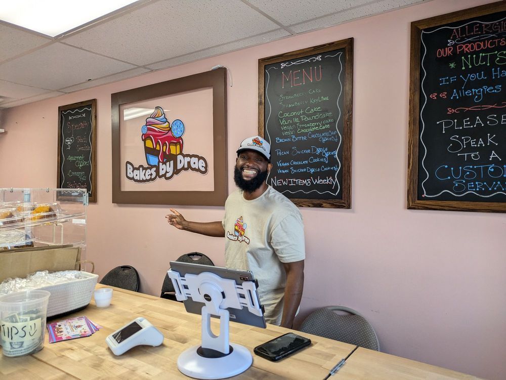 Man in bakery gestures towards a counter with a tablet. Menu boards and bakery logo on pink wall.