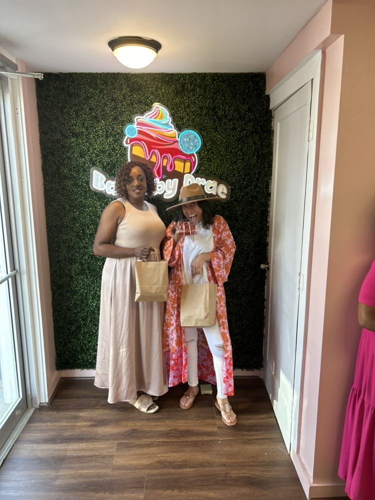 Two women pose by a bakery sign on a green wall. One in a dress holds a bag; the other wears a hat and coat.