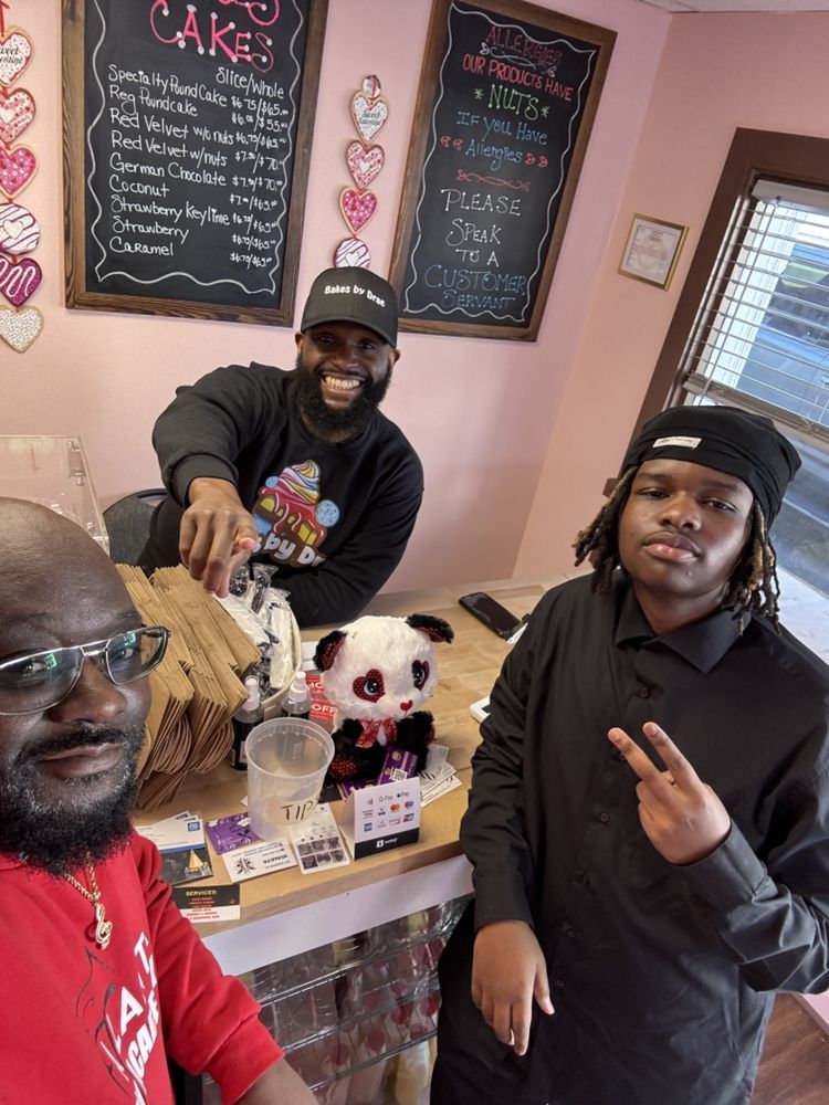 Three people at a pink-walled bakery counter, one smiling, pointing, and two looking at the camera. A panda plush sits nearby.