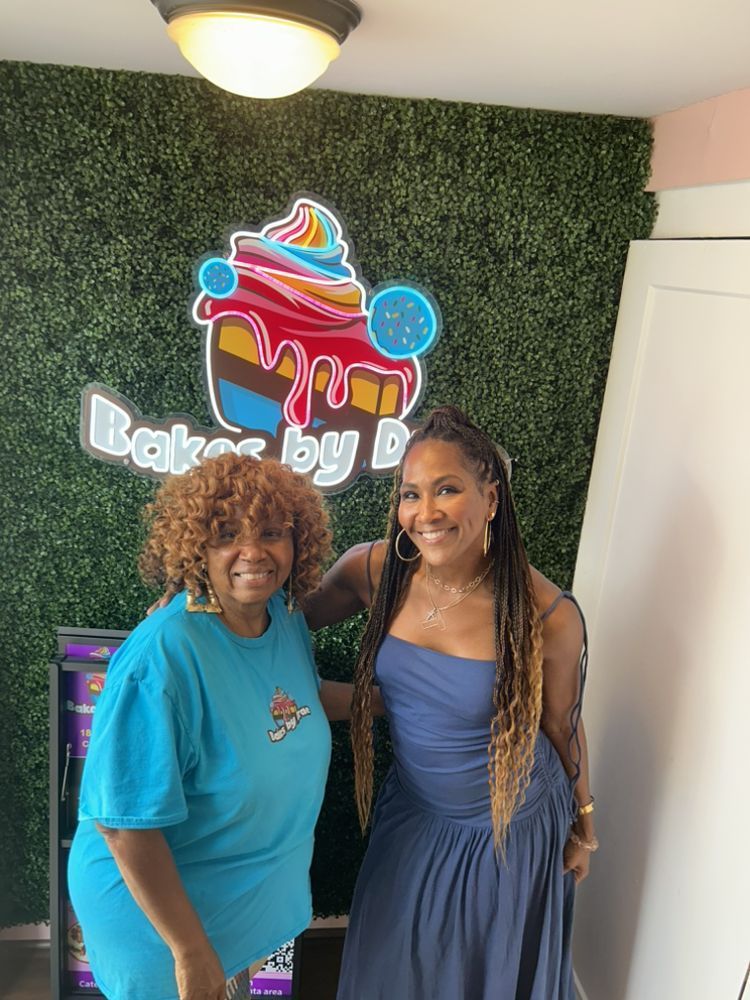 Two women pose in front of a bakery logo. One wears blue, the other a long blue dress. Bakery has a green wall.