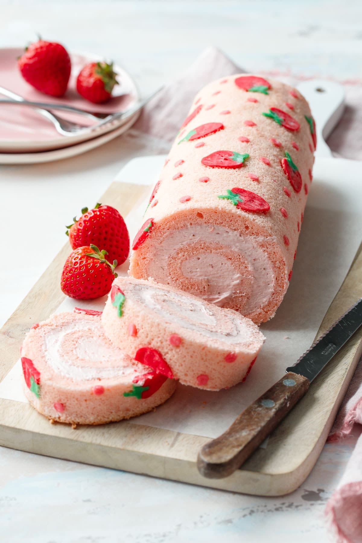 Strawberry roll cake on a wooden board with fresh strawberries and a knife. Pink frosting and decorations.
