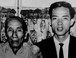 A man in a suit and tie is standing next to an older woman in a black and white photo.