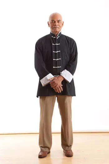 A man in a black jacket and tan pants is standing with his hands folded.
