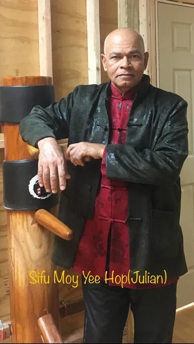 A man in a black jacket is standing next to a wooden dummy.