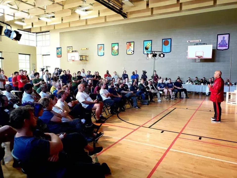 A man in a red jacket is standing in front of a large group of people in a gym.