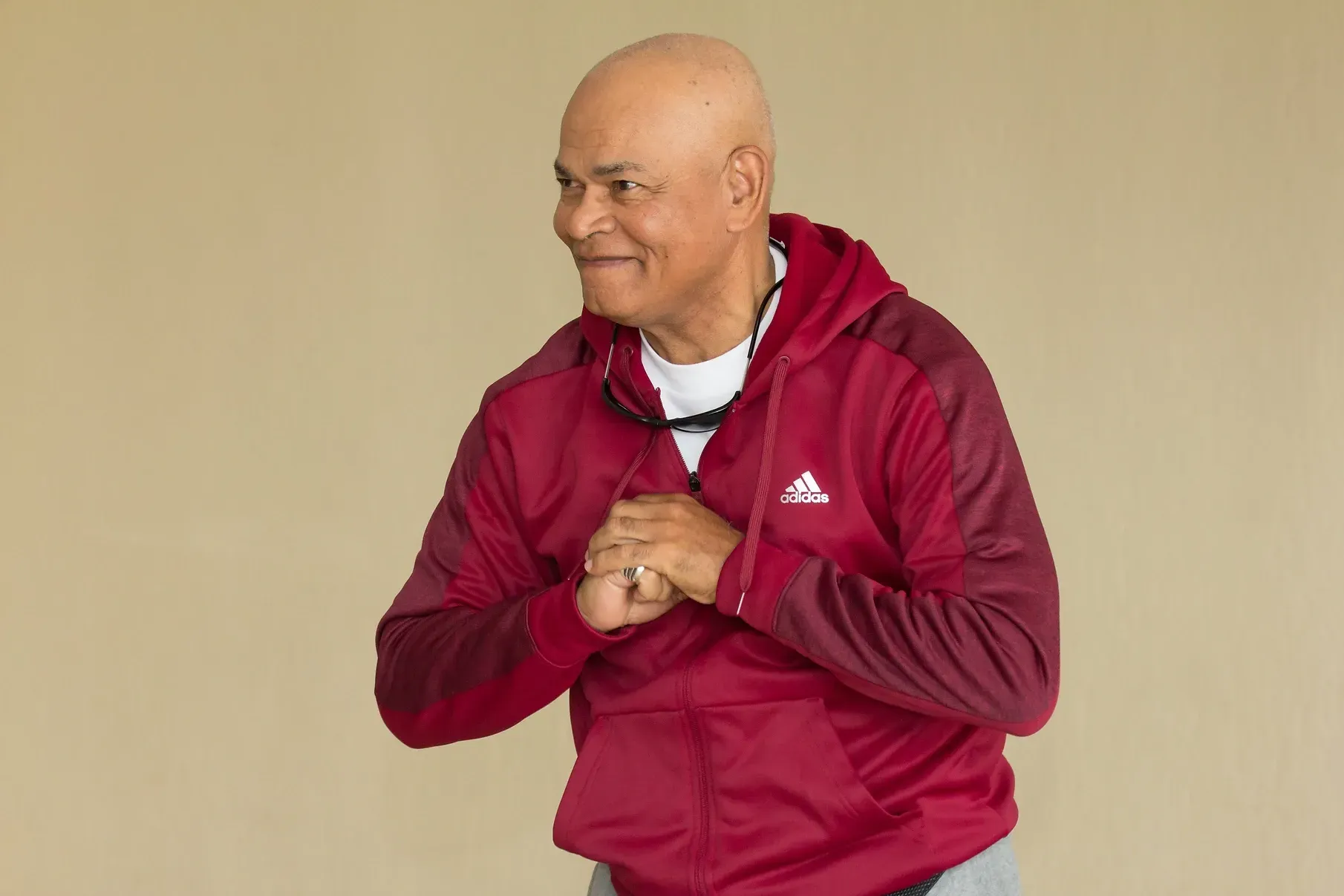 A bald man wearing a maroon adidas jacket is standing with his hands folded.