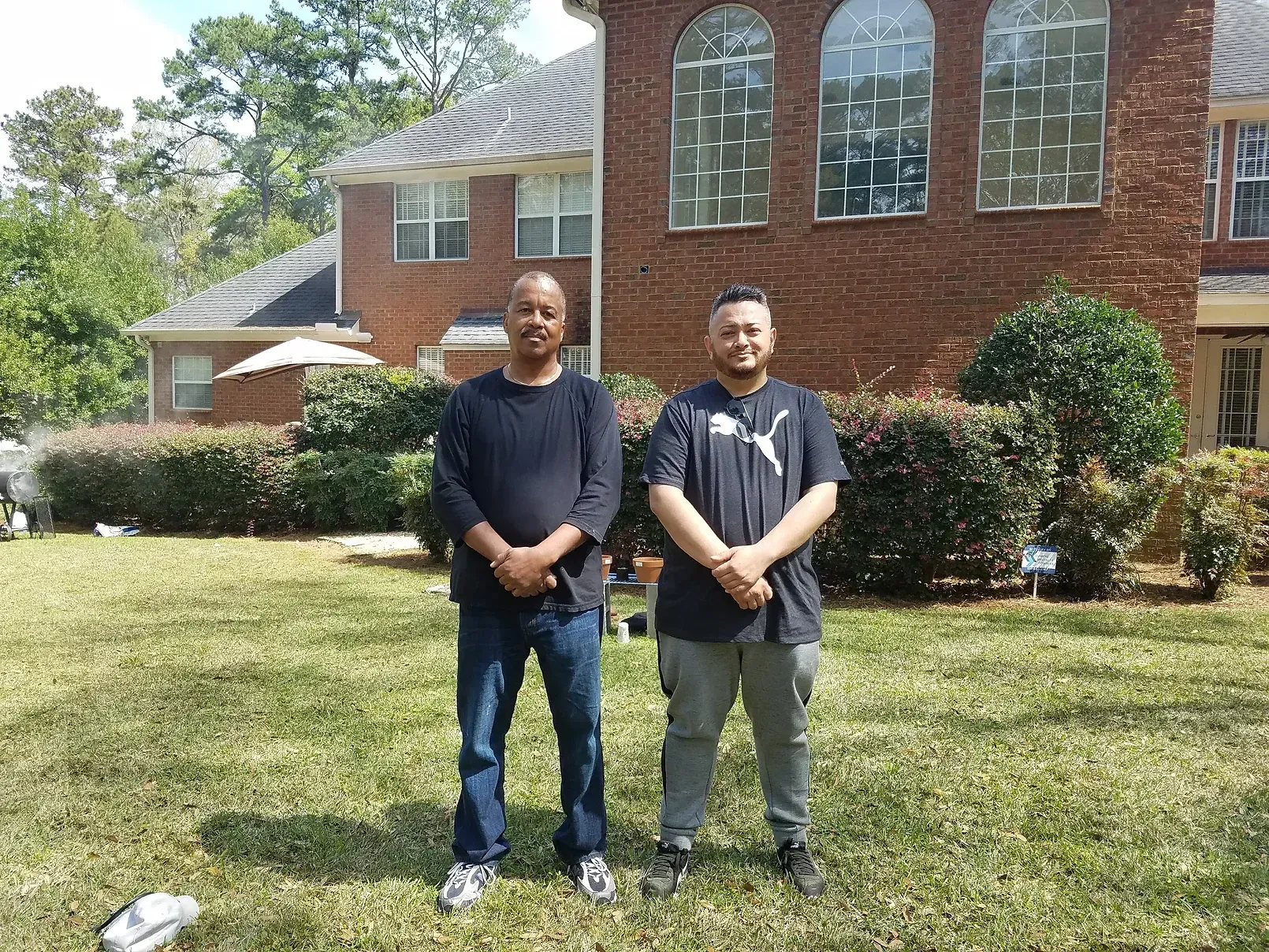 Two men are standing in a grassy field in front of a brick house.