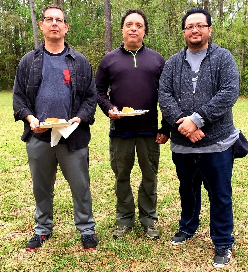 Three men are standing in a grassy field holding plates of food.