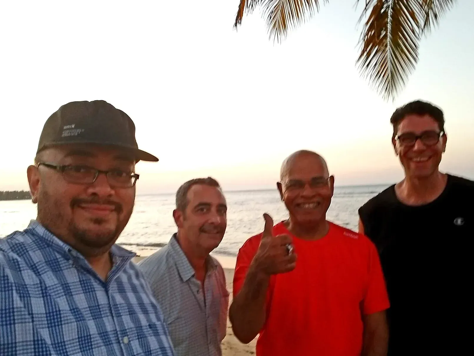 Four men are posing for a picture on the beach and one of them is giving a thumbs up