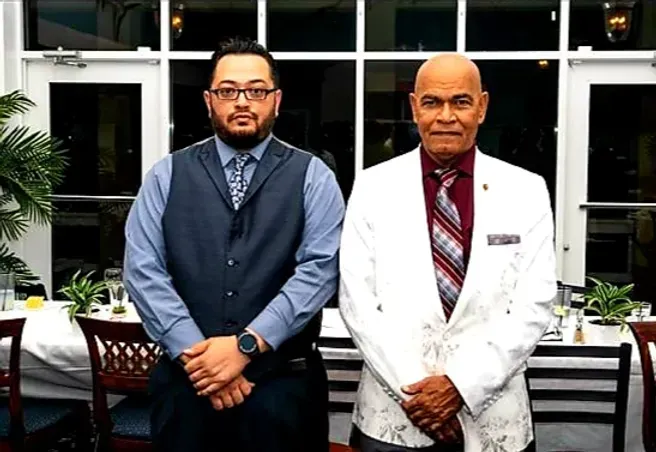 Two men in suits and ties are standing next to each other in front of a window.