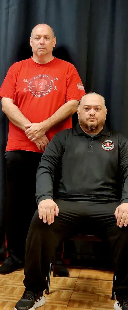 Two men are standing next to each other and one is sitting in a chair.