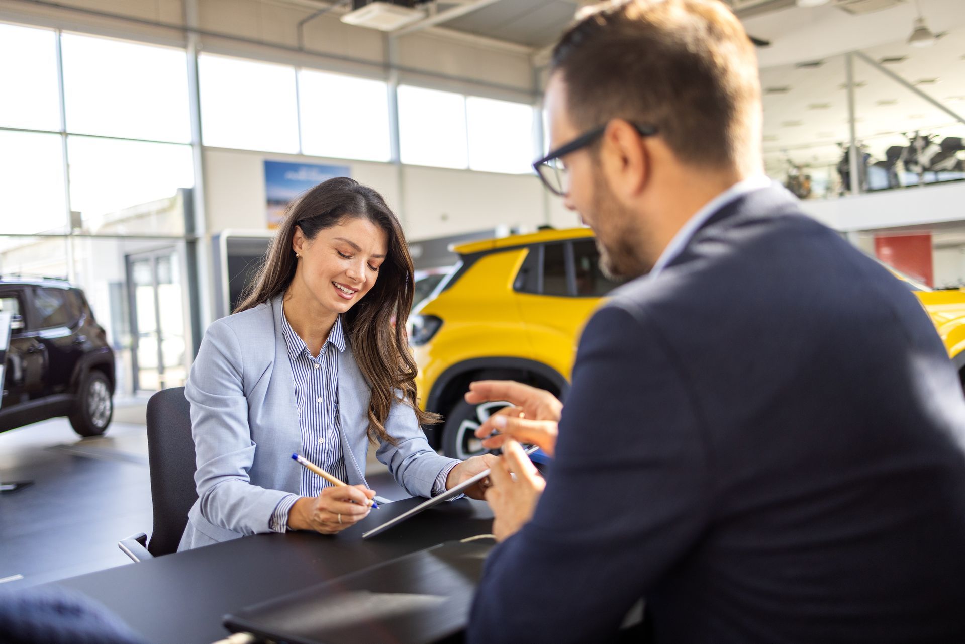Smiling woman signs on a tablet while a man explains her coverage at an auto insurance agency desk. Smiling woman signs on a tablet while a man explains her coverage at an auto insurance agency desk.