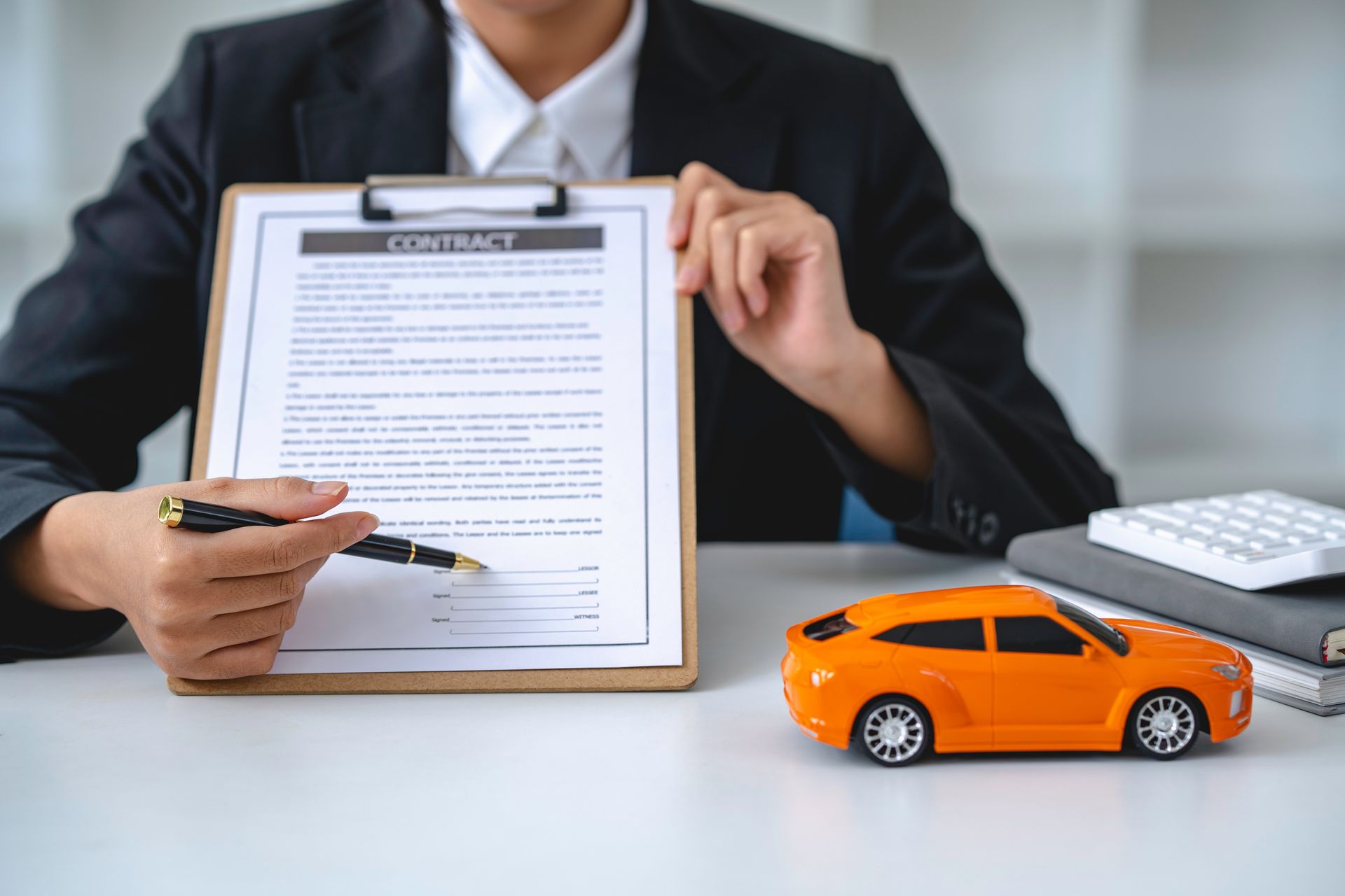 Finger pressing a car insurance start button inside a vehicle. Hand pointing a pen at an auto insurance contract on a clipboard, next to a toy car and calculator.