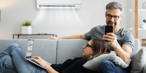 Two people relaxing on a couch, under a working air conditioner. One using a laptop, the other with a phone.