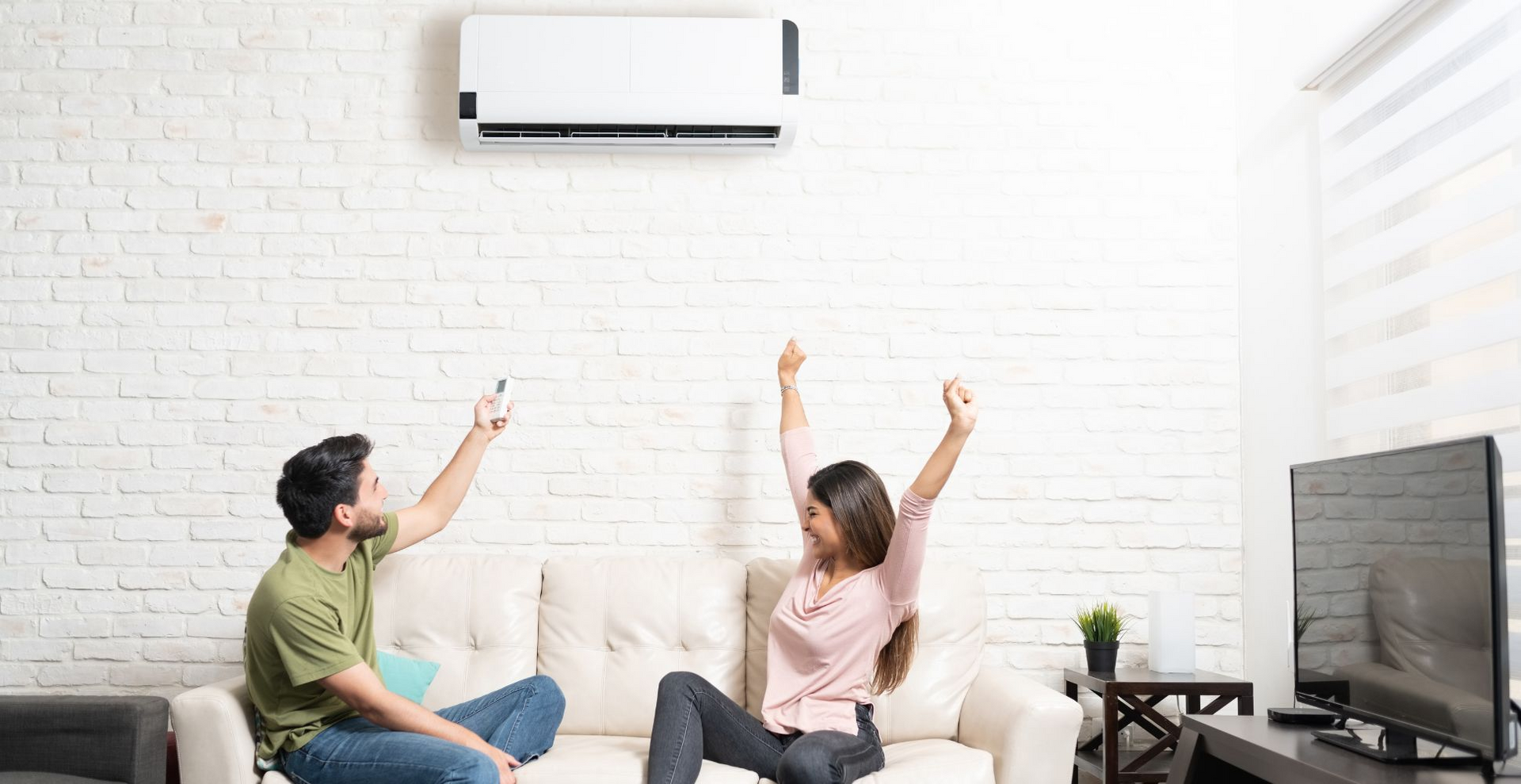 A couple celebrates, adjusting air conditioner with remote on a white brick wall.