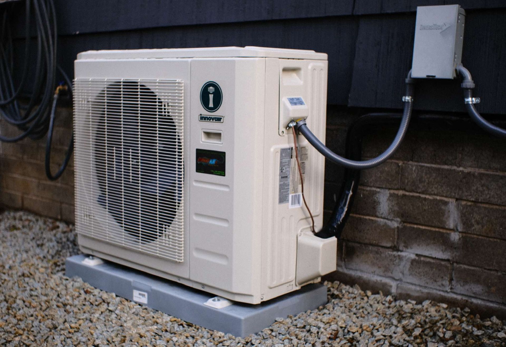 Beige heat pump unit on a gray base, against a dark wall and gravel.