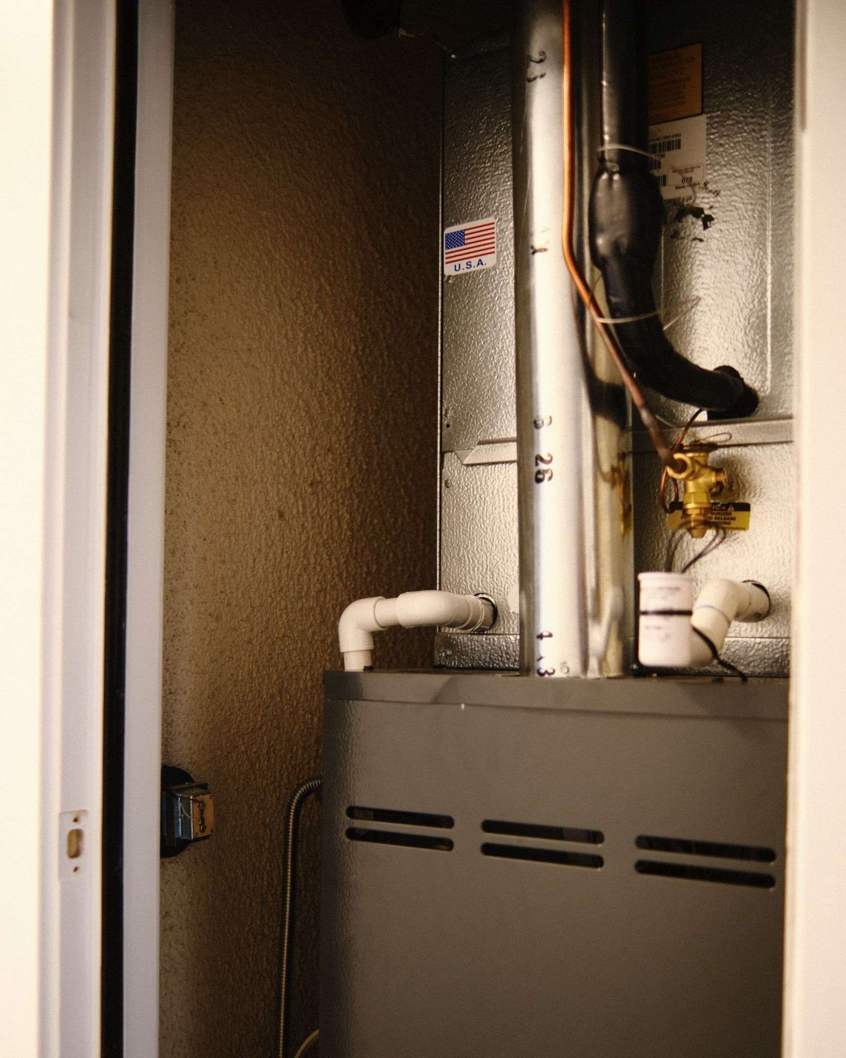 Furnace unit inside a closet with ductwork and pipes. Silver and gray with white pipes.