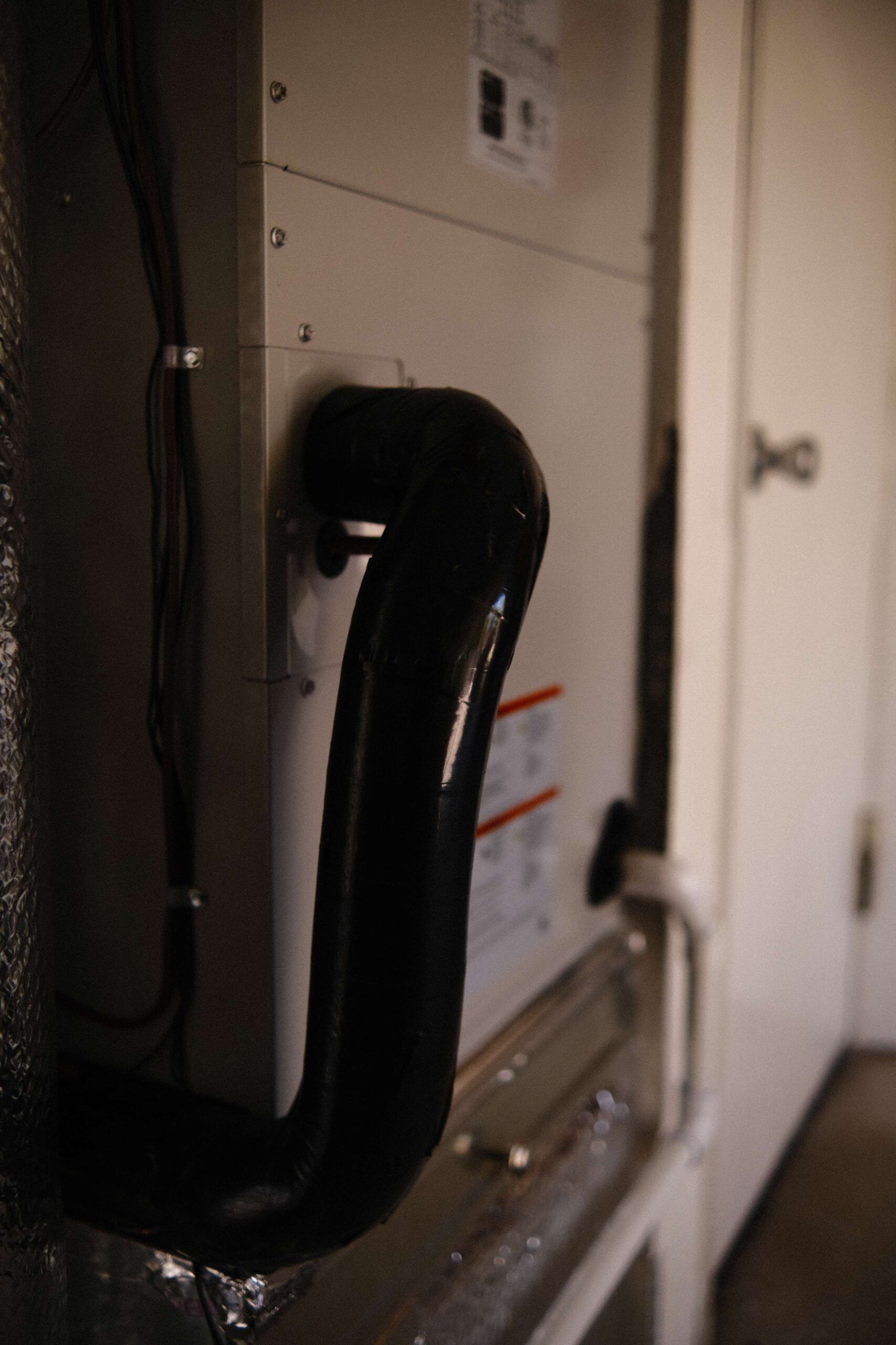 HVAC unit with black pipe exiting, mounted on a wall in a room. Door in the background.