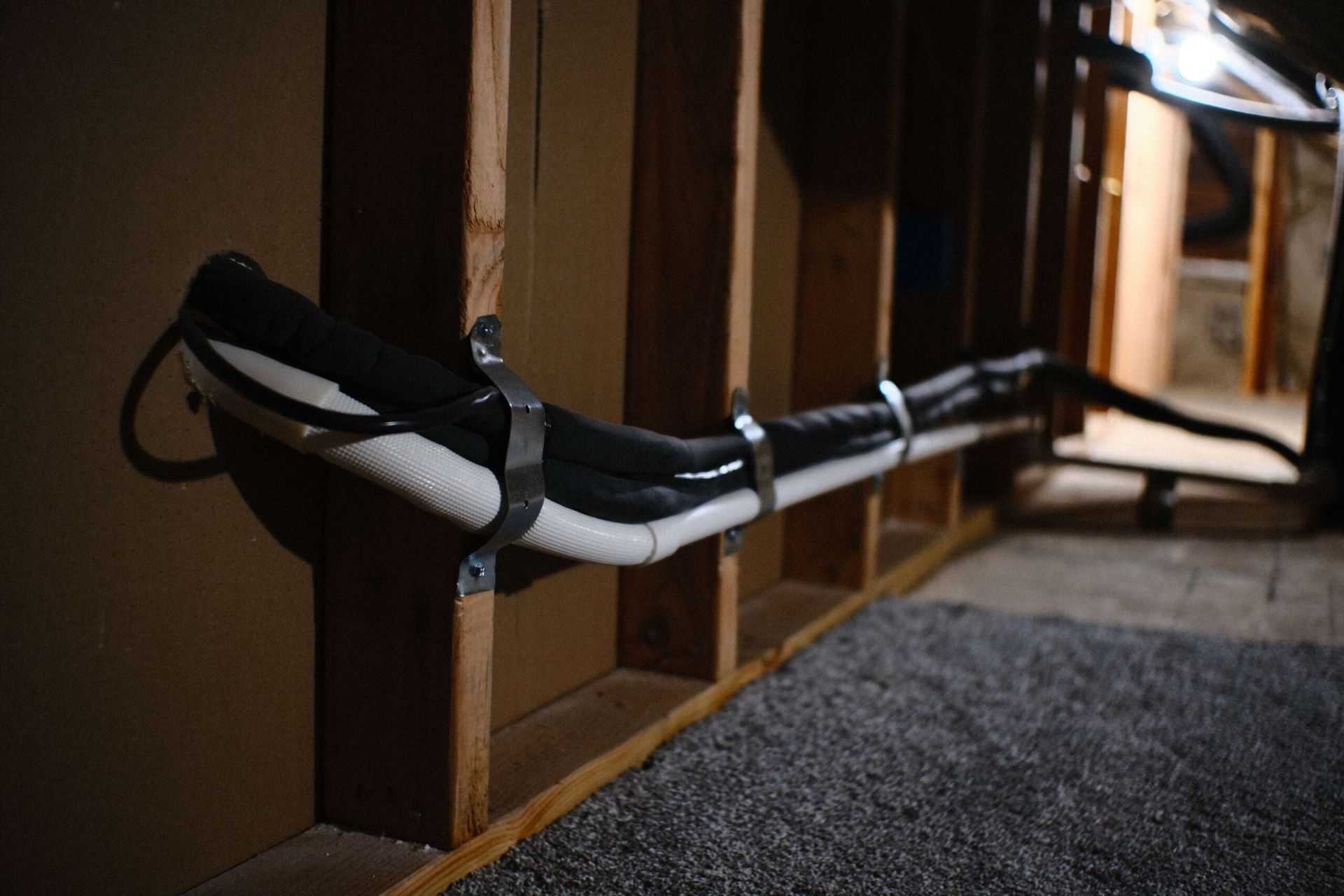 Black and white pipes secured to wooden studs with metal clamps in a dim basement.