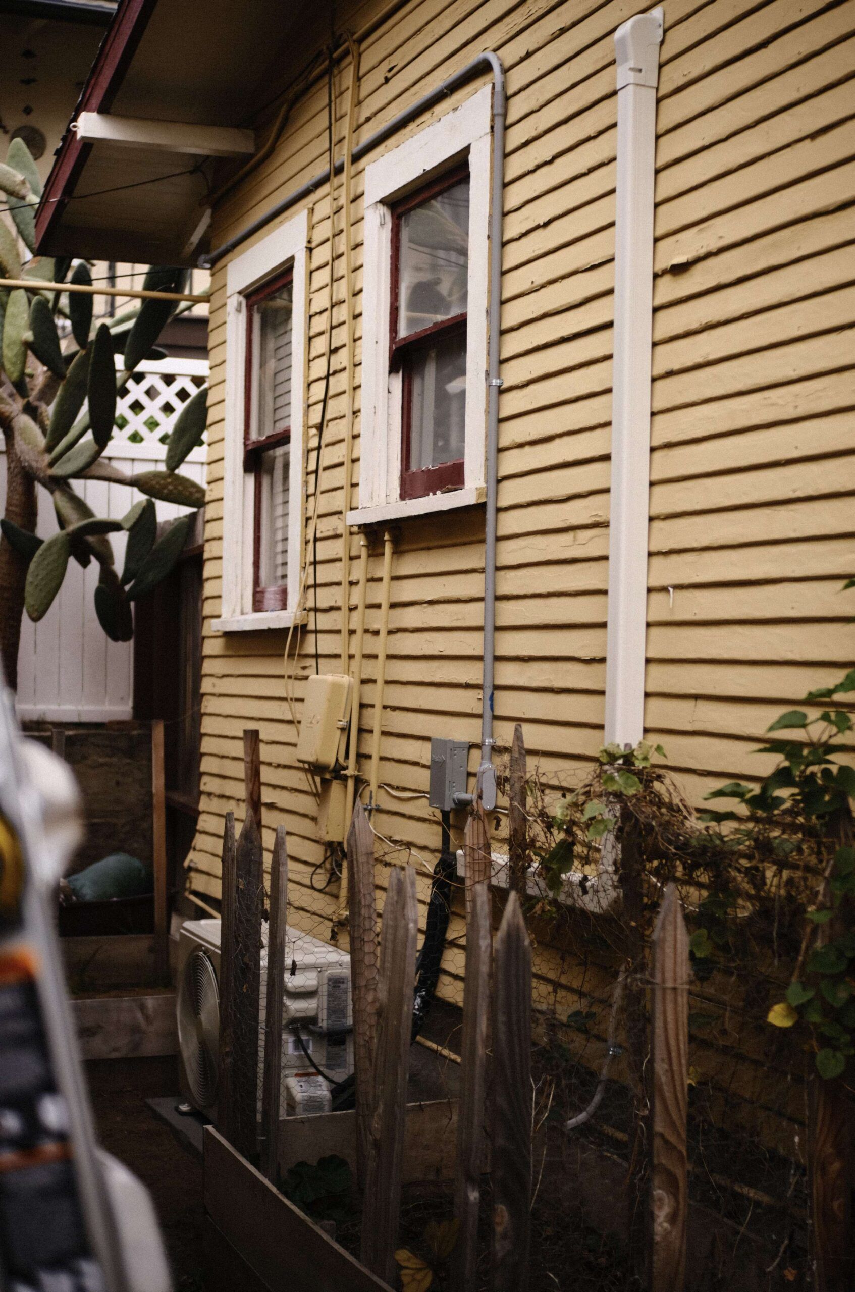Yellow wooden house exterior with two windows, electrical boxes, and a wooden fence.