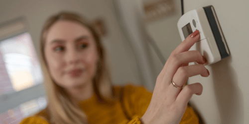 Woman's hand adjusts a thermostat on a white wall. She wears a gold ring and orange sweater, face out of focus.